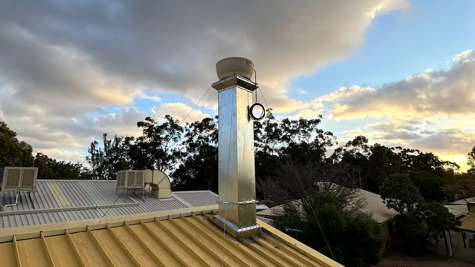 A fabricated galvanised flue riser through a Colorbond roof, silhouetted against a sunset sky