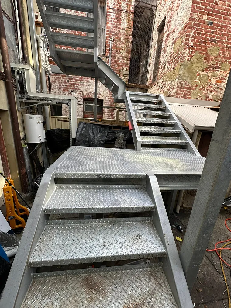 A galvanised steel switchback stair with checker-plate treads installed against a weathered heritage brick wall