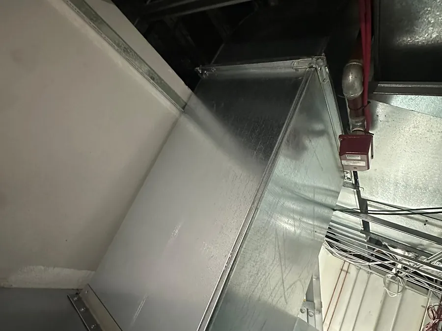 A large galvanised sheet-metal duct transition with riveted flanges installed above a ceiling