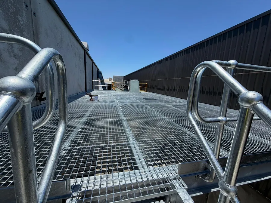 A galvanised steel access platform with mesh decking and tubular handrails installed between an industrial shed and a precast concrete wall